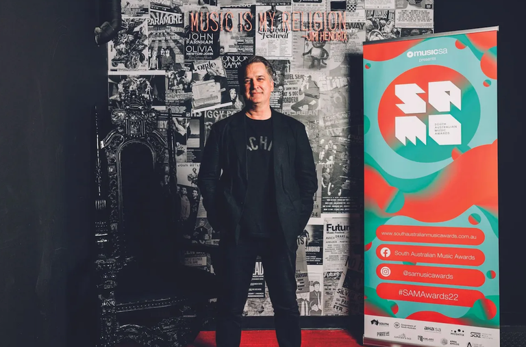 Joe Hay posing next to a South Australian Music Awards banner on a red carpet with gig posters and a throne in the background for a SAM Awards photo.